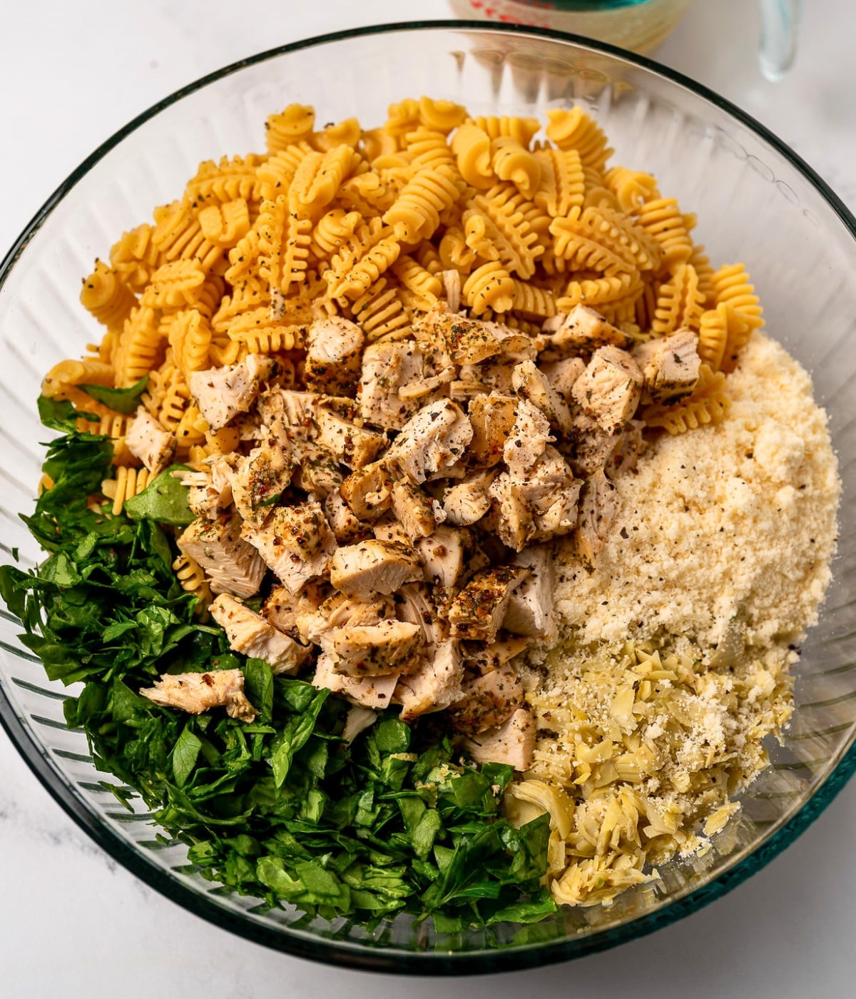 air fried and chopped chicken breast in a mixing bowl with cooked protein pasta, artichoke hearts, leafy greens, and parmesan cheese