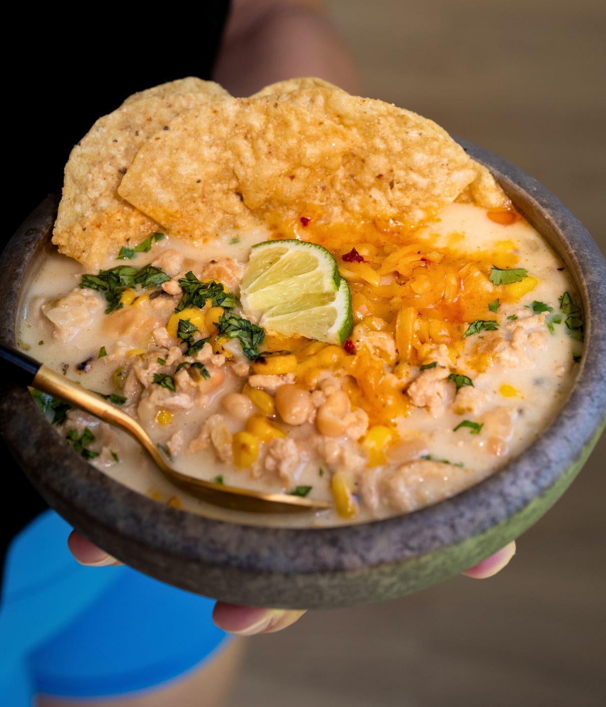 holding a bowl of white chicken chili topped with cheddar cheese, tortilla chips, and lime