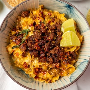 coconut curry noodles in a bowl topped with crispy ground beef crumbles, lime wedges, and chili crisp