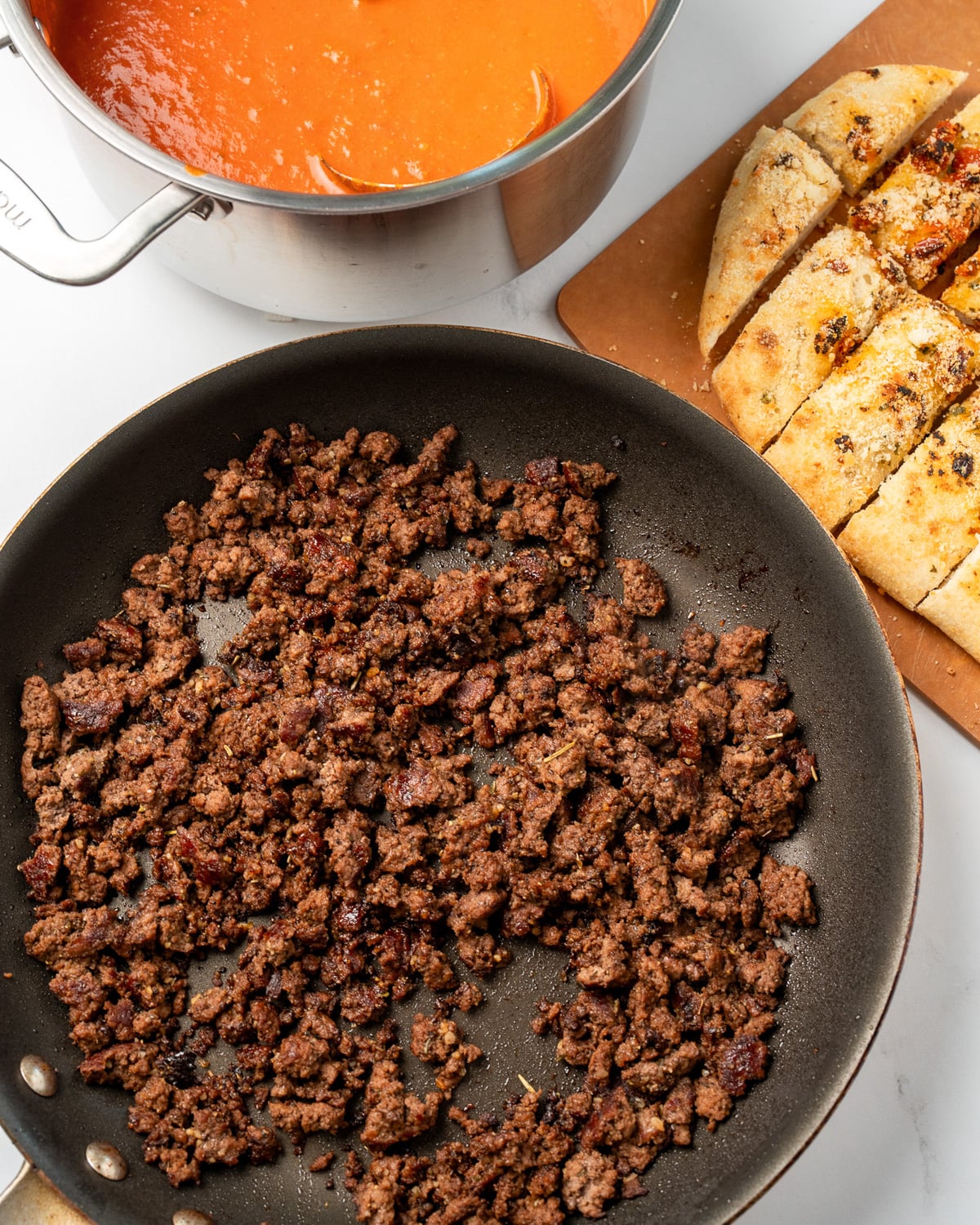 cooked balsamic ground beef crumbles in a pan next to warmed tomato soup in a pot and sliced focaccia on a cutting board