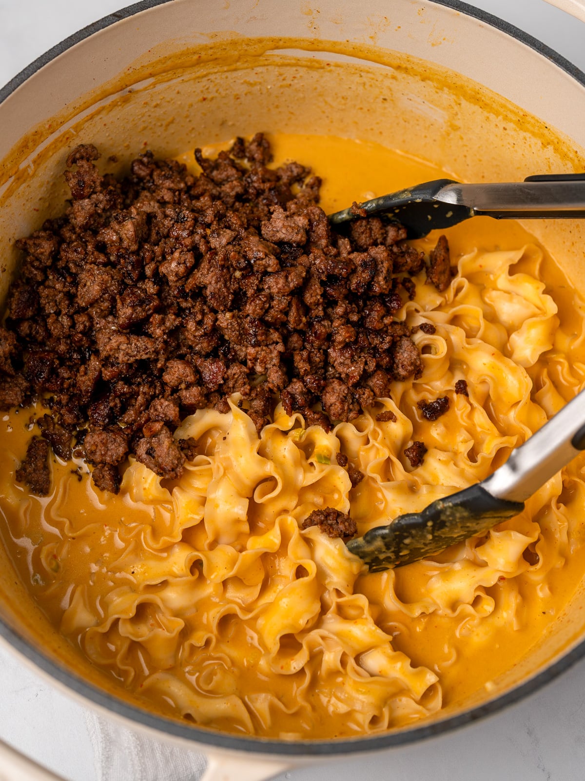 crispy ground beef crumbles in a pot with creamy pumpkin noodles and tongs