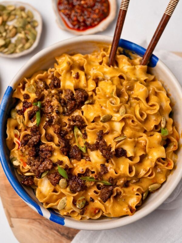 chopsticks in a bowl of creamy pumpkin noodles topped with crispy ground beef crumbles, roasted pumpkin seeds, and sliced green onion